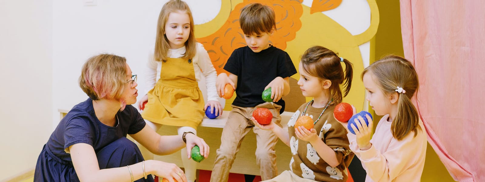 A woman interacts with a group of four children that are holding objects.