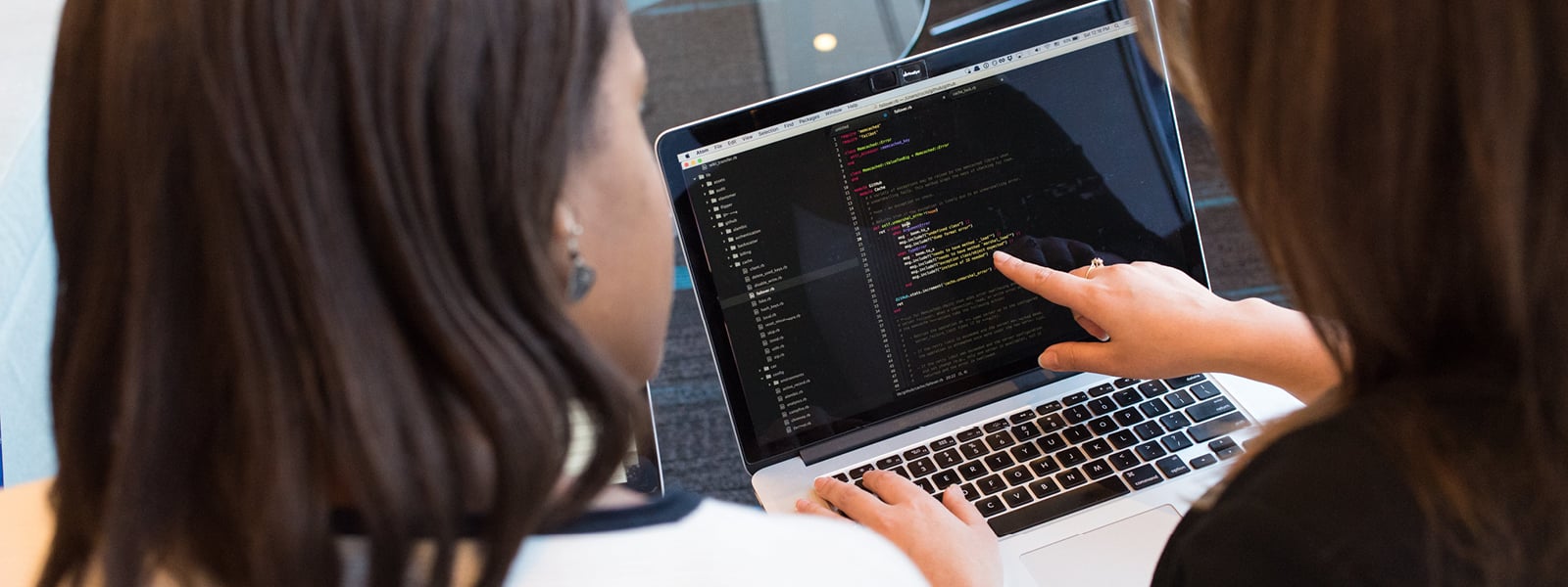 A student points to code on a computer screen while another student watches.