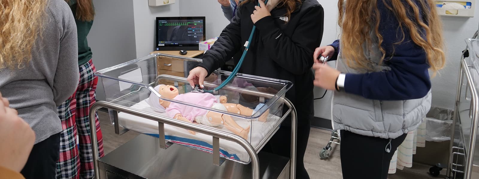 A student using a stethoscope on a dummy infant while other students watch.