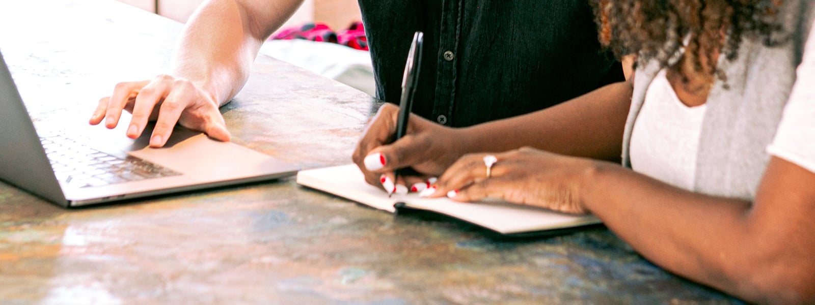 A woman writes in a notebook as a man uses a laptop. 