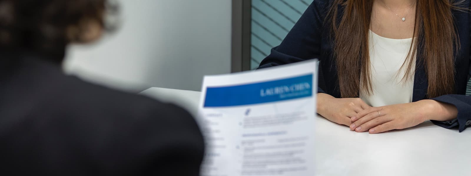 A businesswoman sits at a table while another person holds a piece of paper. 