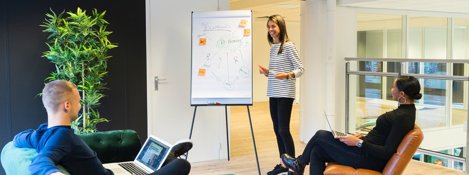 A woman stands at a display board with writing and sticky notes on it as two other people listen.