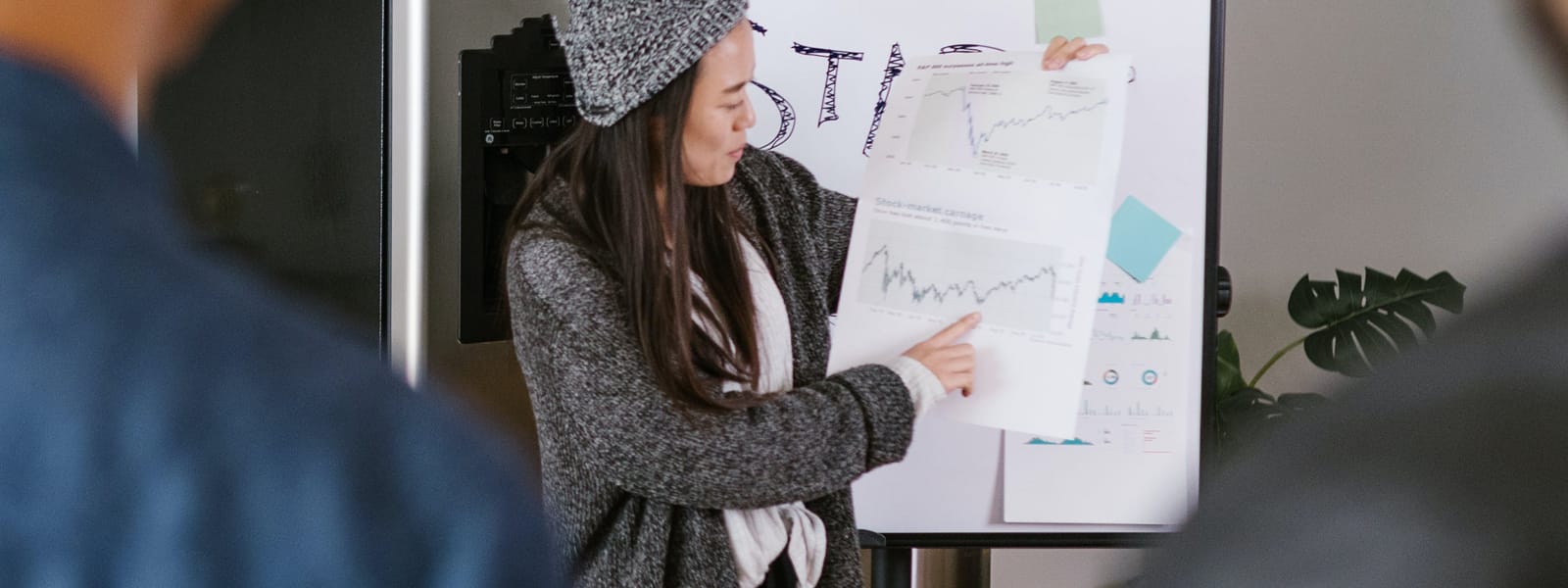 A woman points to a line chart on a paper in front of a presentation board while two people watch.