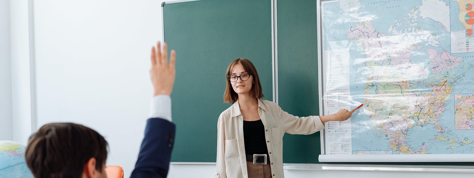 A professor pointing at a map in the front of a classroom as a students raises his hand. 